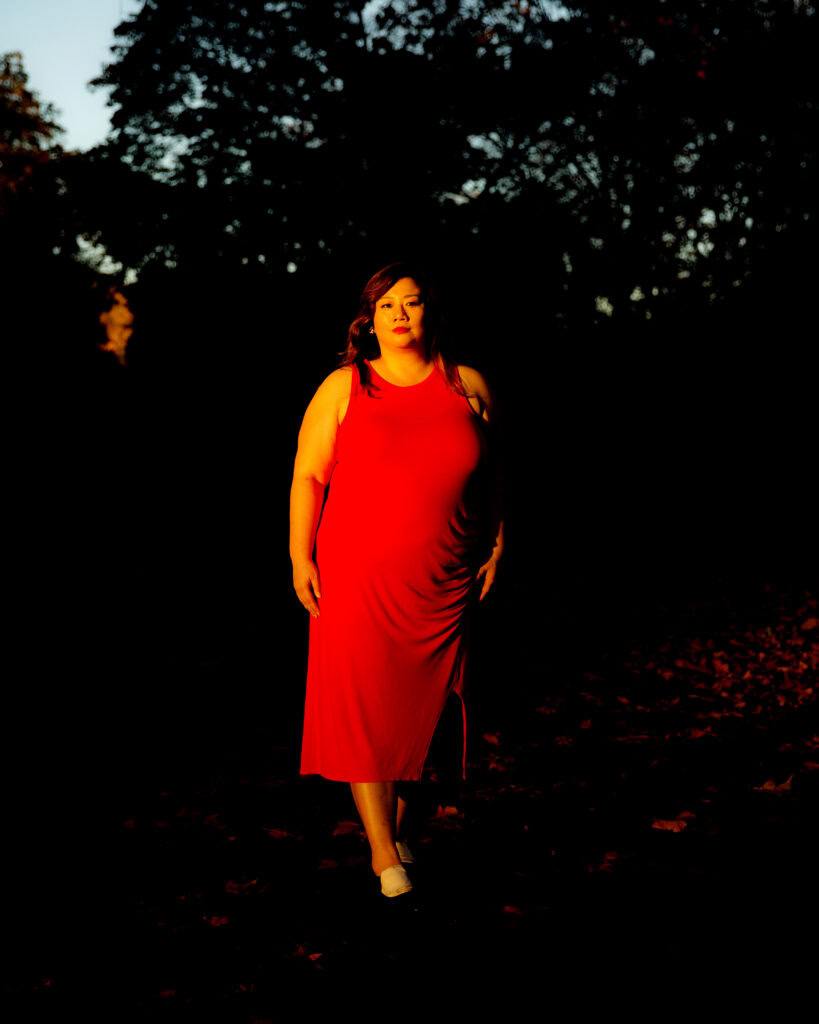 Vanessa Chan, photographed by Mary Inhea Kang. Chan wears a red dress and stands in front of a dark background of trees