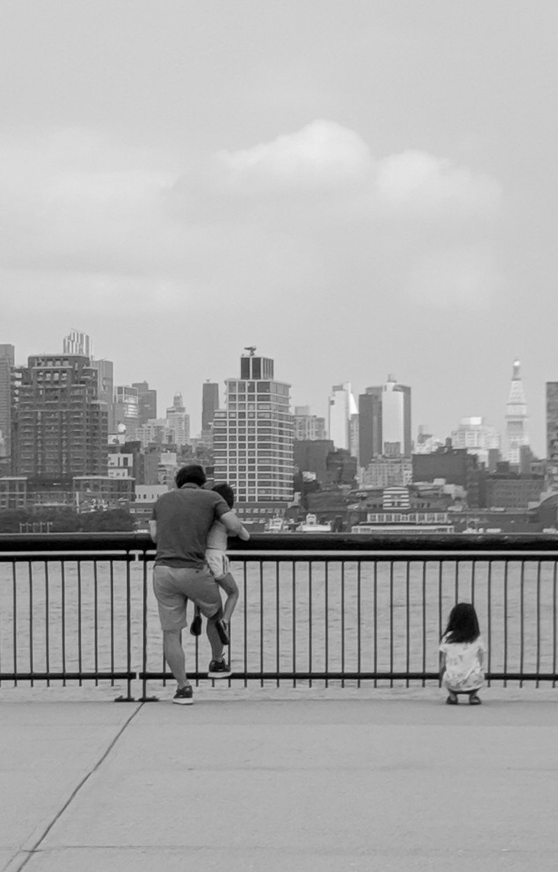 Black and white photograph of man and children looking over the river at NYC skyline