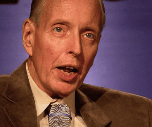 Close up of a man in a brown suit jacket speaking at a microphone