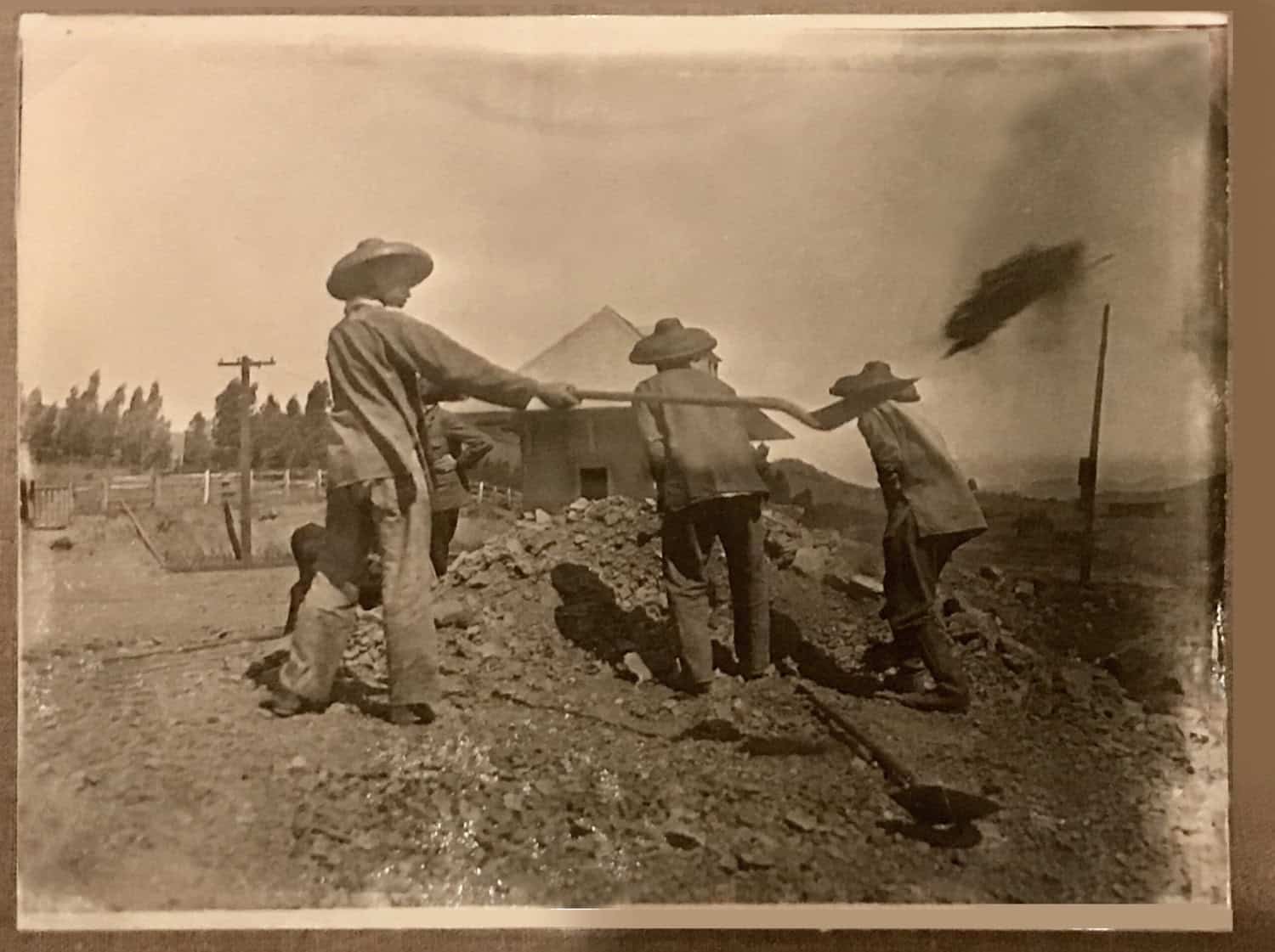 Historical grainy photograph of three men in wide brimmed hats digging a mound of earth with shovels. They are wearing loose fitting clothes. There are trees and a small building in the background.