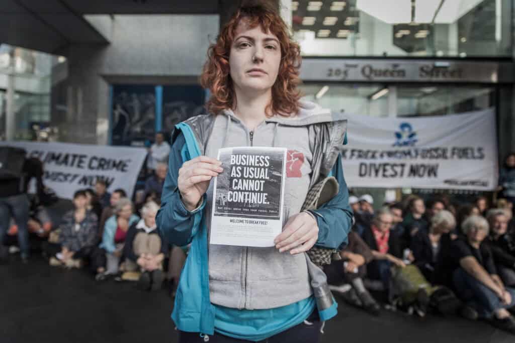Woman with red curly hair holds a sign that reads Business As Usual Cannot Continue. In the background is a large group of people sitting and standing to form a protest against fossil fuels.