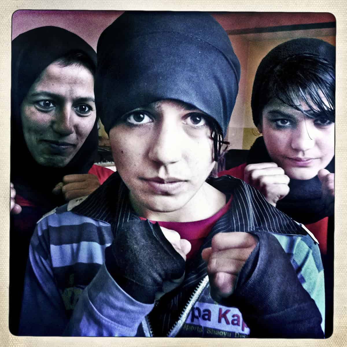 Two young female boxers with raised fists face the camera. An older woman looks on.