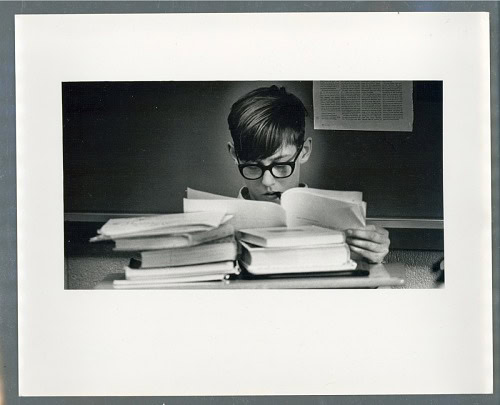 Black and white photograph of teen boy reading a large stack