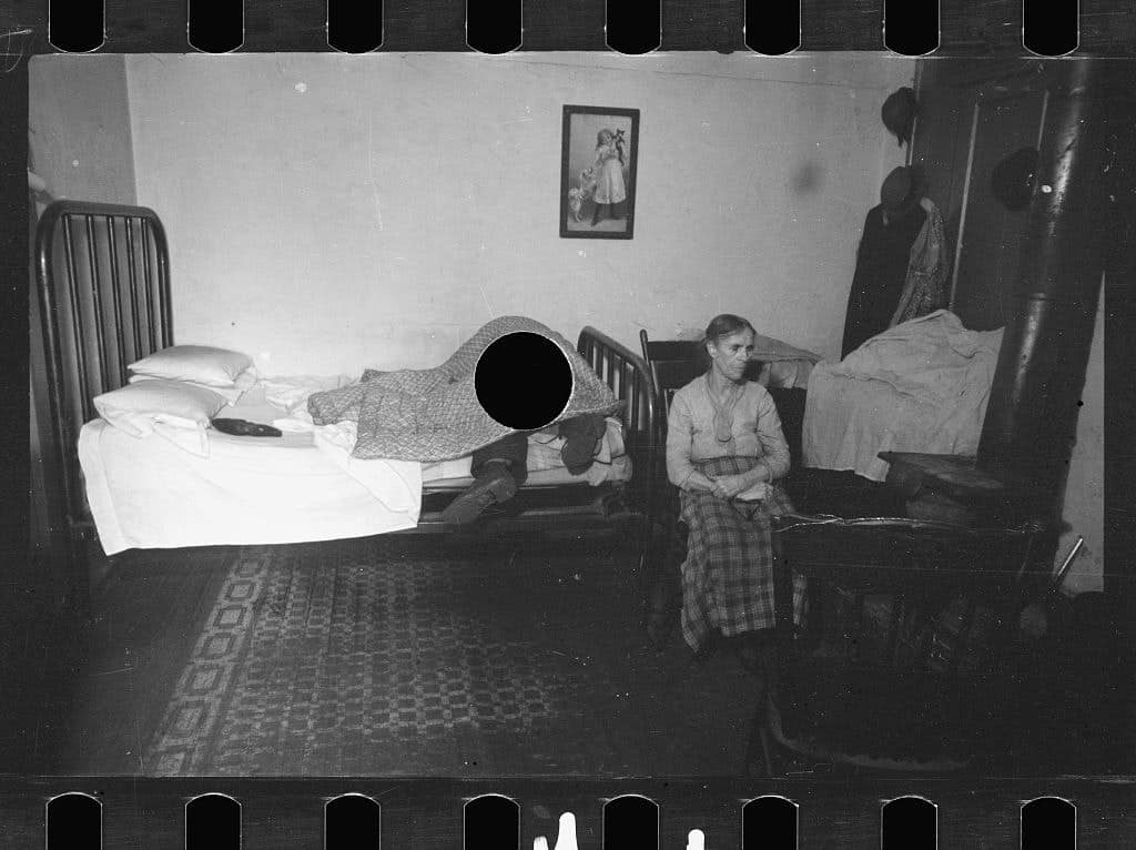 Black and white image photograph of woman sitting next to bed on which a man's boots are visible
