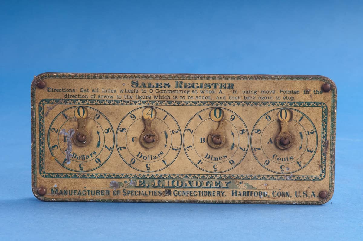 This small device has a tin case painted yellow and green on top and red around the edge. The top of the case has four wheels drawn on it, each of which has the digits from 1 to 9 drawn around the edge. The digits go clockwise for the first and third circles (marked “Cents” and “Dollars” and counterclockwise for the second and fourth circles (marked “Dimes” and “Dollars”). At the top of each circle, at the zero position, is a window that reveals a rotating disc below. The discs are rotated using thumbscrews that protrude from the back of the instrument. An arm on top of each circle points to a digit on the wheel. The discs advance when they are rotated in the direction of increasing digits and remain fixed when the arrows are moved back to zero.