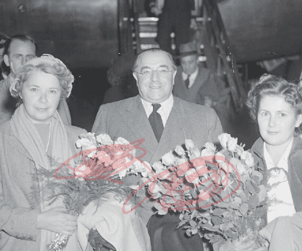 Black and white photo of older man in suit getting of airplane flanked by women carrying bouquets