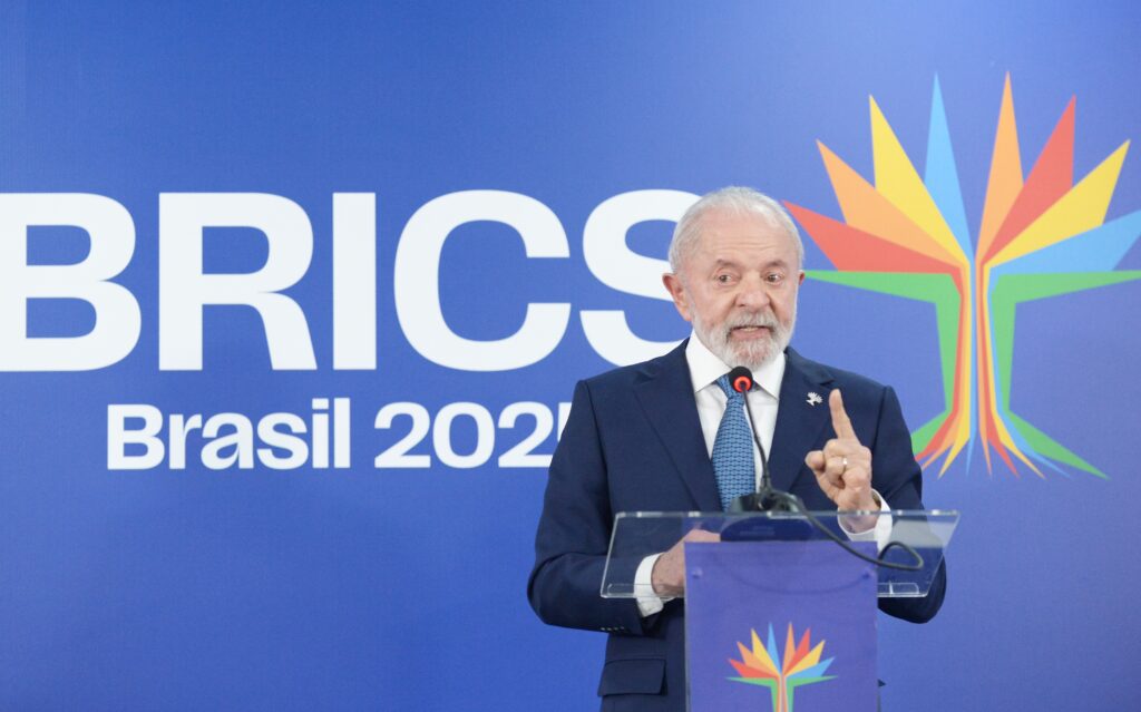 Man in blue suit with white beard stands at a conference podium, holding up his index finger as he speaks