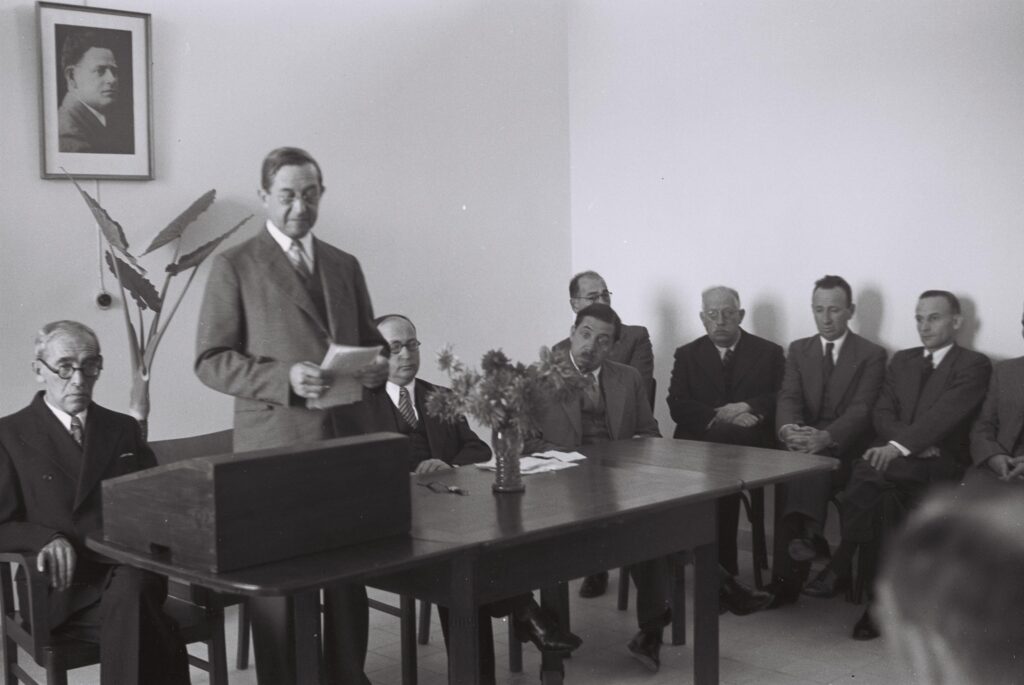 Black and white photograph of a suited man reading a speech. The man stands in front of a table. Seven men in suits sit behind him.