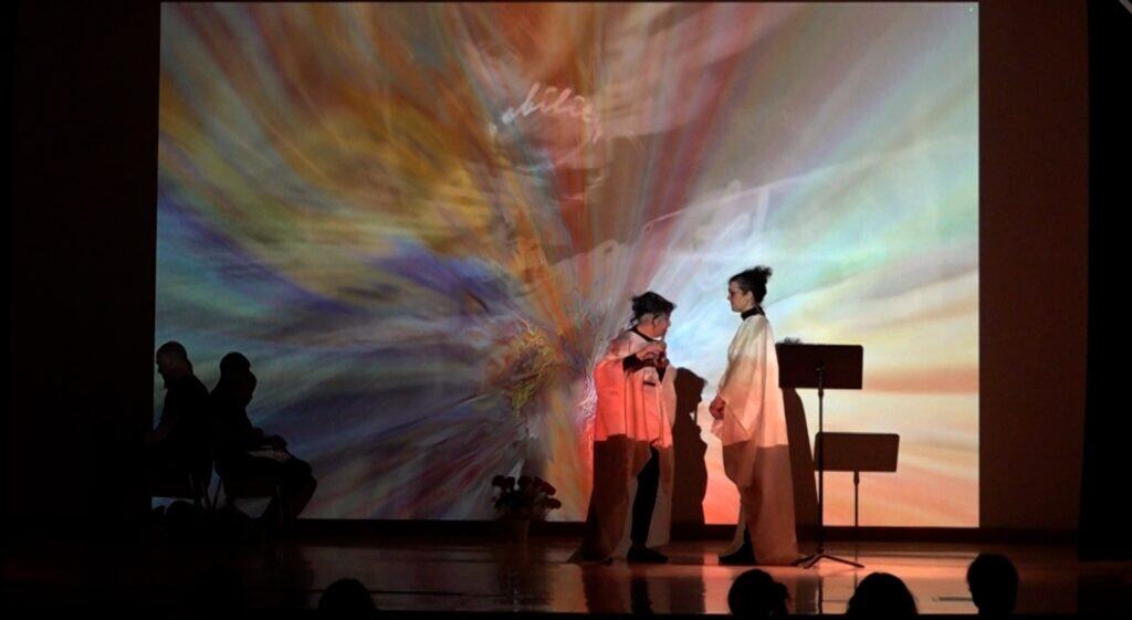 Two figures in white robe stand on stage in swirling colored light