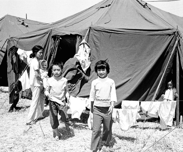 Vietnamese refugee families, including women and children, walk past rows of tents in a temporary resettlement camp in the United States after the fall of Saigon, 1975.