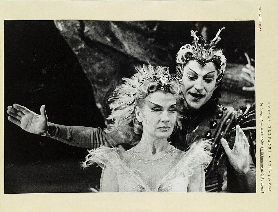 Black and white photograph of a woman in a white dress and elaborate surrealist crown; a dark clad winged figure leans over her shoulder