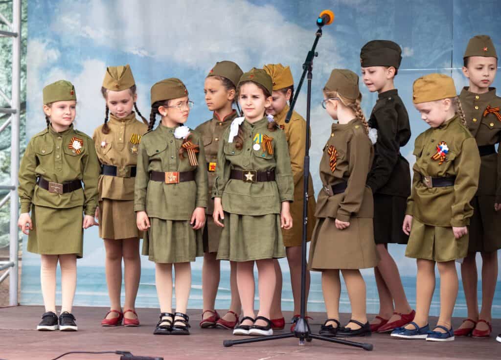 A group of children in military uniforms stand on a stage. The children are wearing green uniforms and are standing in a line