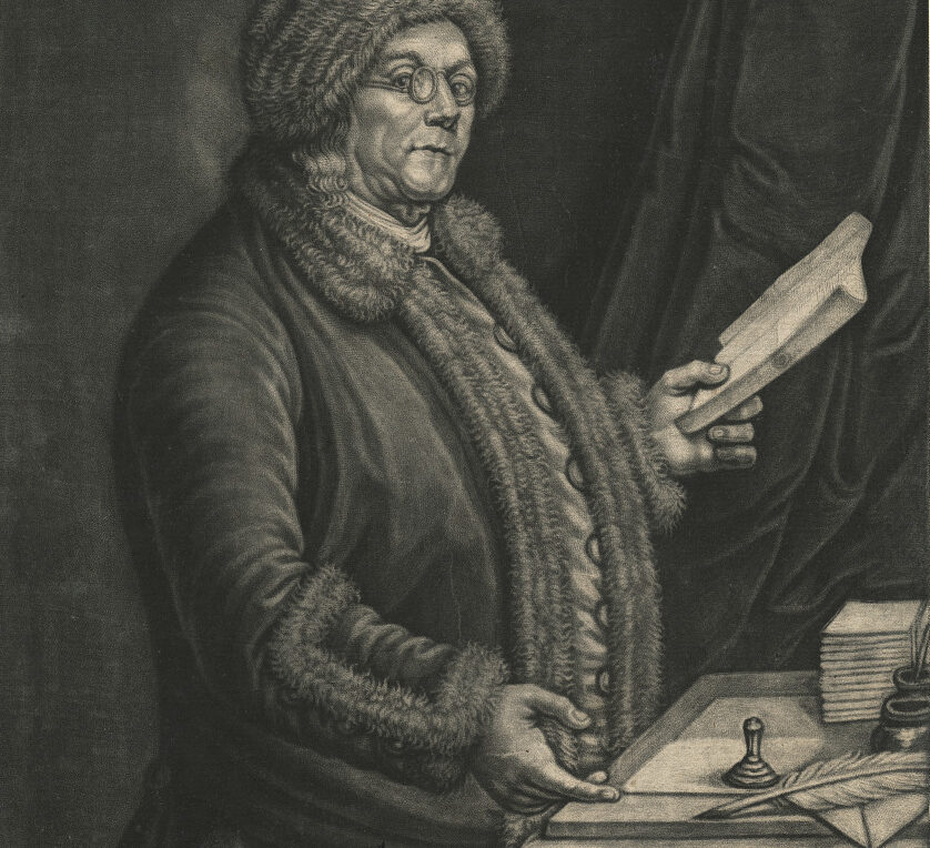 Black and white mezzotint on paper of man in spectacles and fur hat holding a book in his left hand. His right hand gestures to a quill and piece of paper on the desk in front of him