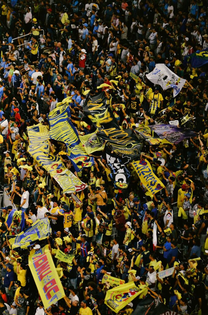 A large crowd of soccer fans fills a stadium section, waving yellow and blue flags and banners with the Mexican Club América emblem in celebration.