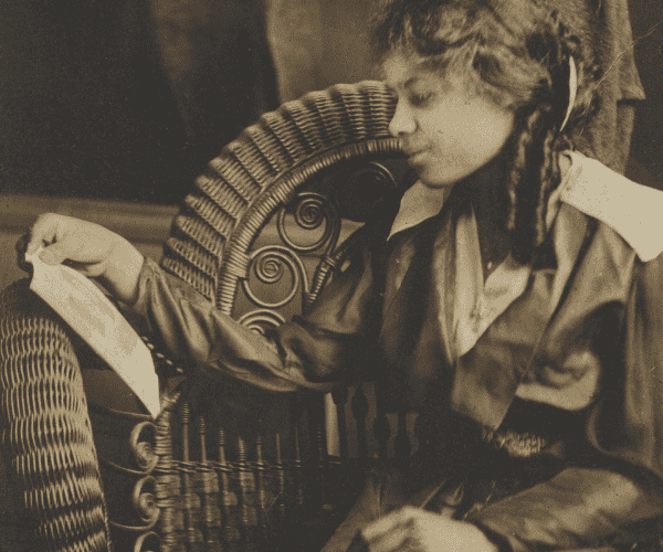 A black-and-white photograph of a young girl sitting on a wicker chair. The young girl is wearing a dark color, loose fitting dress with a light color neckline. She reads from a piece of paper in her left hand. The photograph has a brown mat frame and backing.