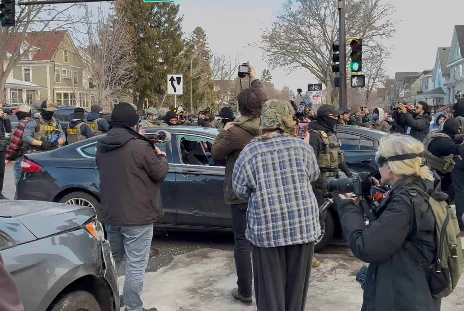 People holding phones and cameras stand around a black sedan with smashed windows; masked ICE agents stand around the car