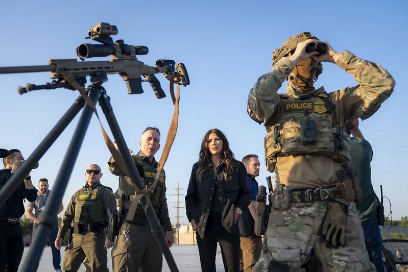 Secretary of Homeland Security Kristi Noem stands between U.S. Immigration and Customs Enforcement officers in camo wear. There is a rifle and scope on a tripod in front of them