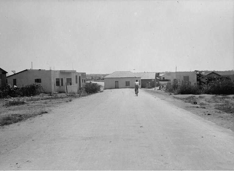 A man rides a bicycle down a dirt road towards a small house built in the middle of the road.