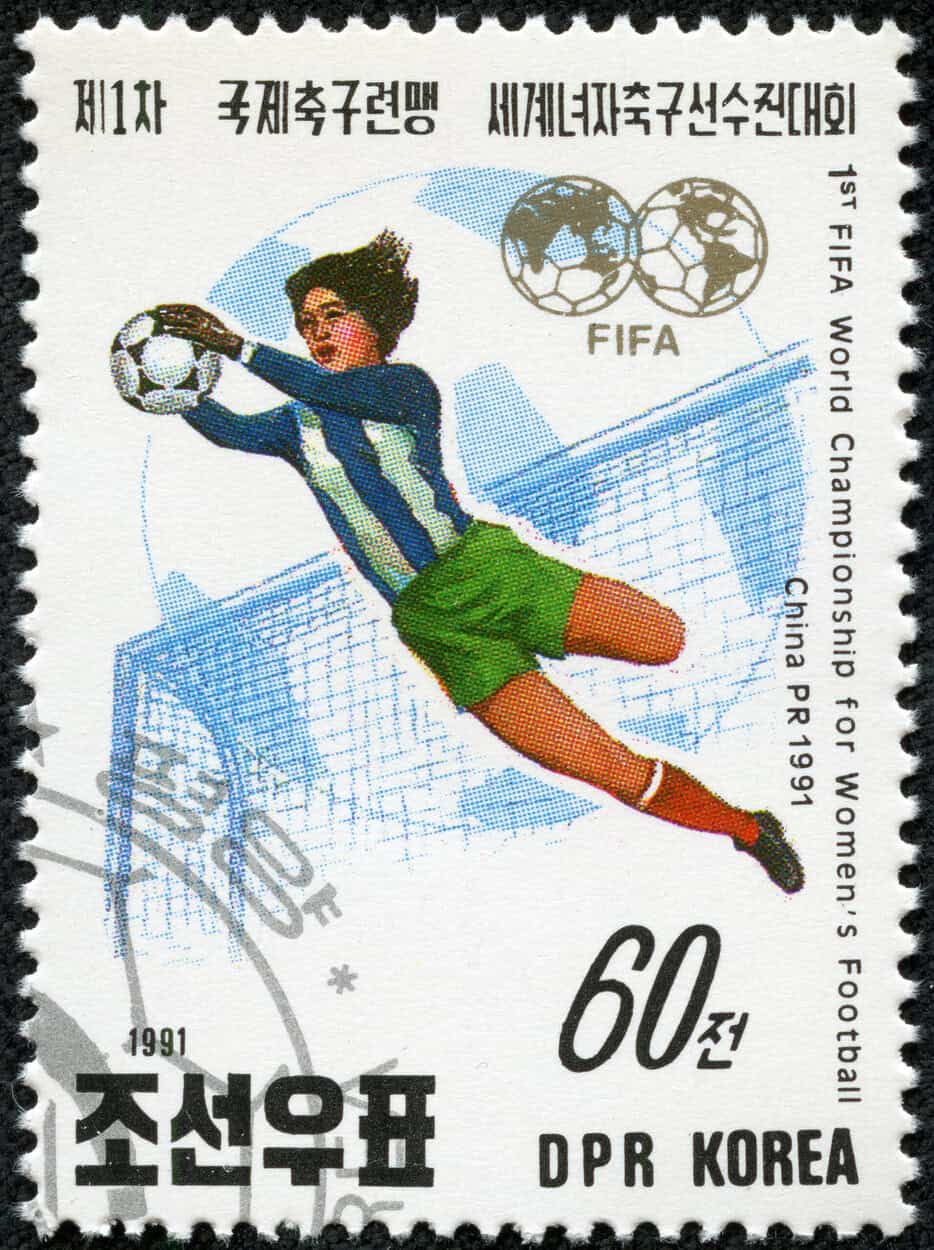 A vintage North Korean stamp depicts a female goalie wearing a blue-striped jersey and green shorts leaping to catch a football