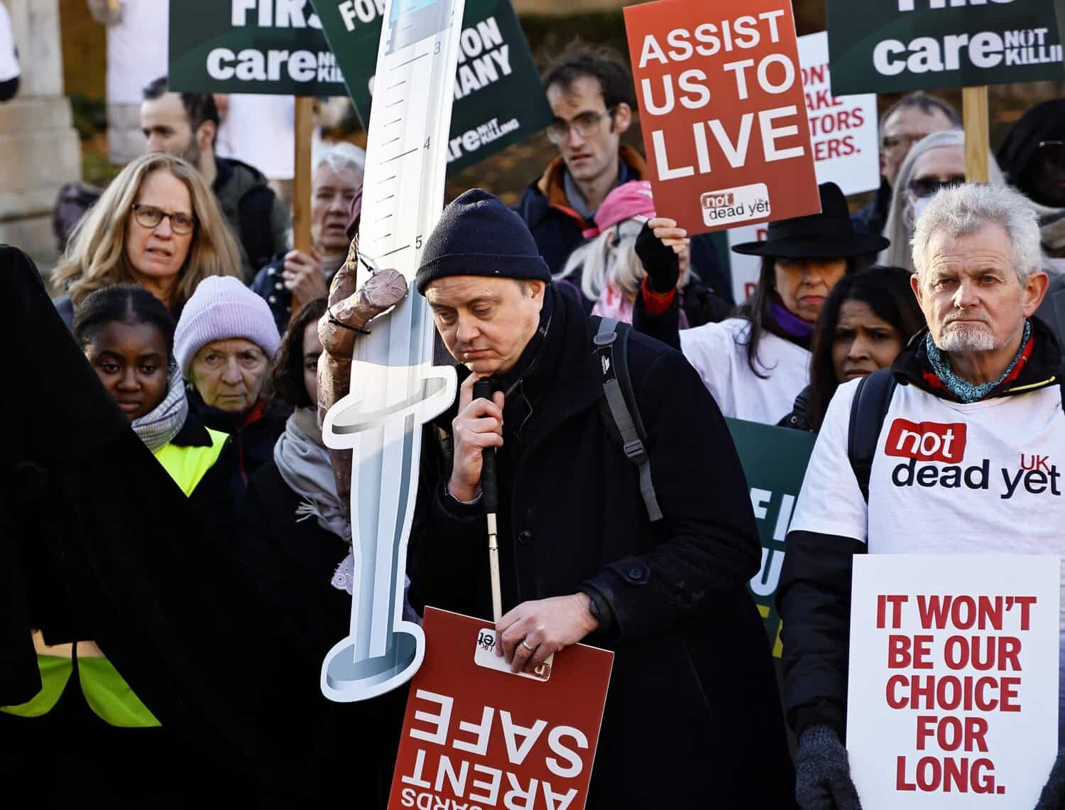 A crowd of English protestors gather holding signs that read ‘Assist us to live,’ and ‘It won’t be our choice for long.’ One man holds a microphone and stands next to a cut out of a giant syringe.