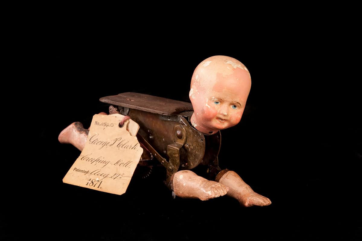 Photograph with black background of crawling white baby doll with realistic head and limbs and wooden mechanical body