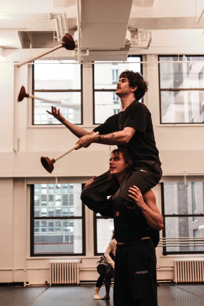 Two dancers in black shirts and dark slacks. One is on the other’s shoulders, juggling three plungers. 