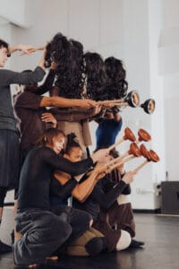 Seven dancers posed in two rows, with three squatting and holding up red plungers, and four standing hanging brunette wigs on sticks and wielding two black plungers.