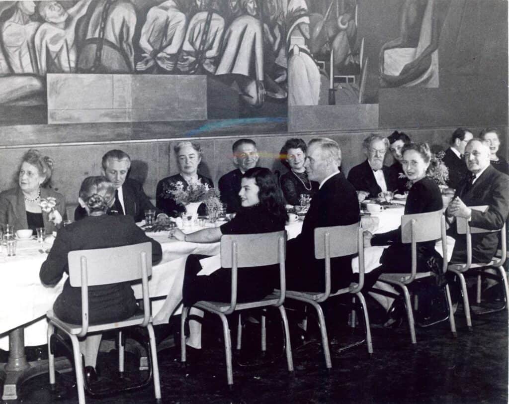 Black and white photo of men and women in 1940s dress seated around a banquet table in hard-back chairs beneath a mural