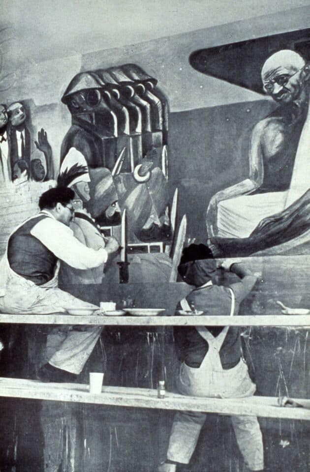 A black and white photograph of José Clemente Orozco and his assistant, Lois Wilcox, working on his mural, “Struggle in the Orient,” in which images of British soldiers wearing gas masks are marching toward Mahatma Gandhi.