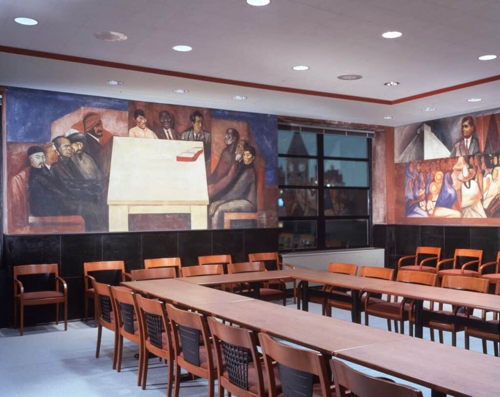 Photograph of wooden classroom chair and table in front of a large fresco showing men of diverse races and ethnicities seated around a table