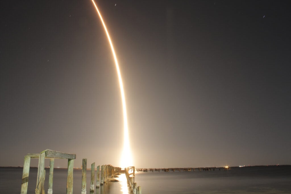 A rocket launch lights up the night sky, foregrounded by a deteriorating wooden dock.