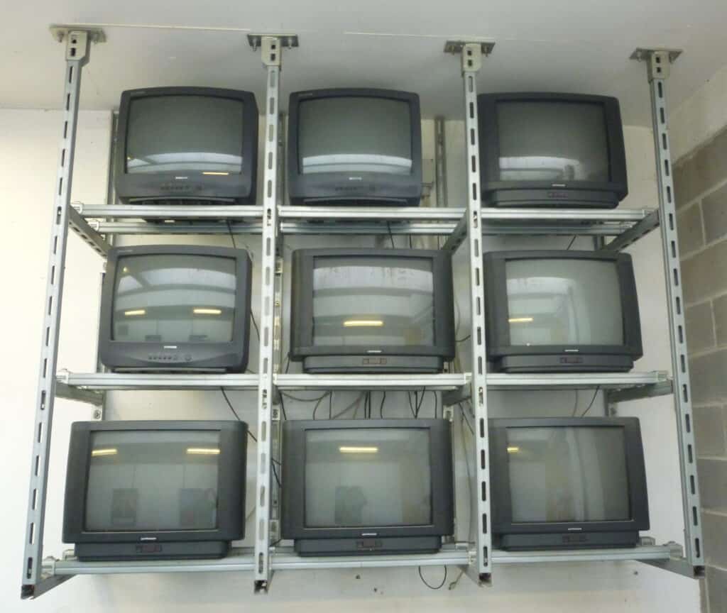 A photograph of a metal rack suspended from the ceiling, holding nine CRT televisions in a three by three orientation.