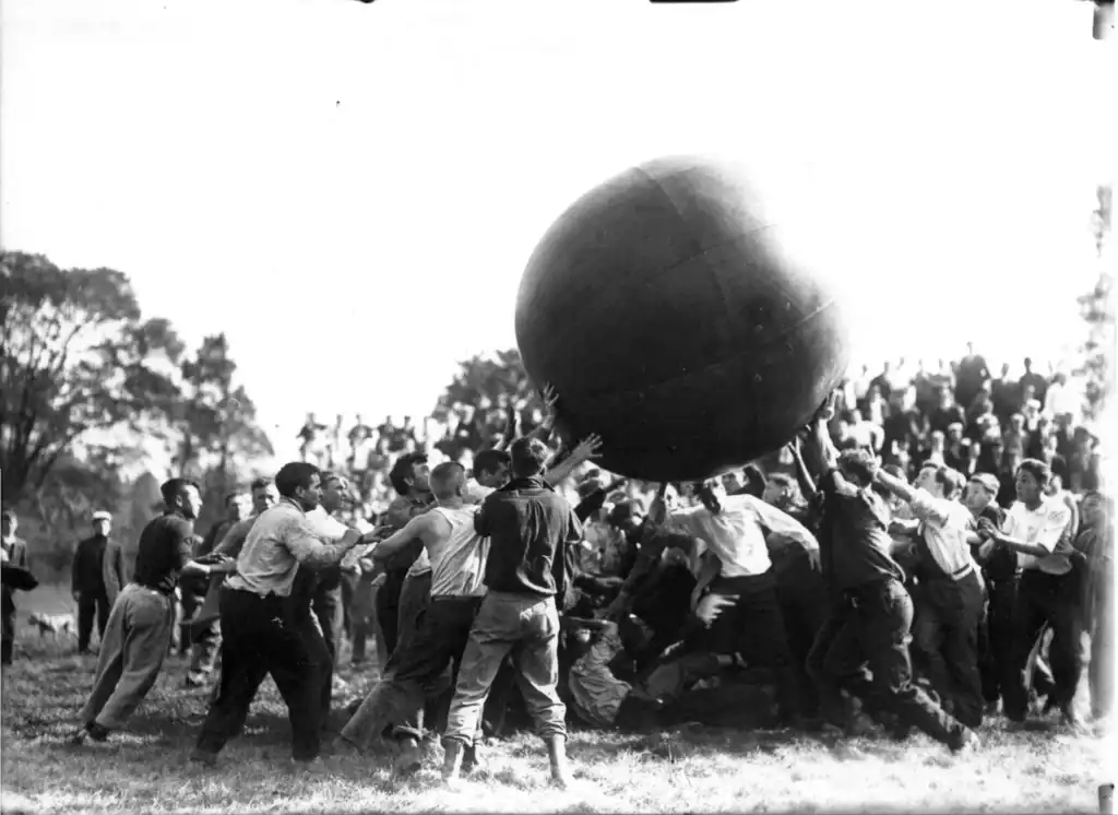 Black and white photograph of crowd of young men competing to hold up a giant ball