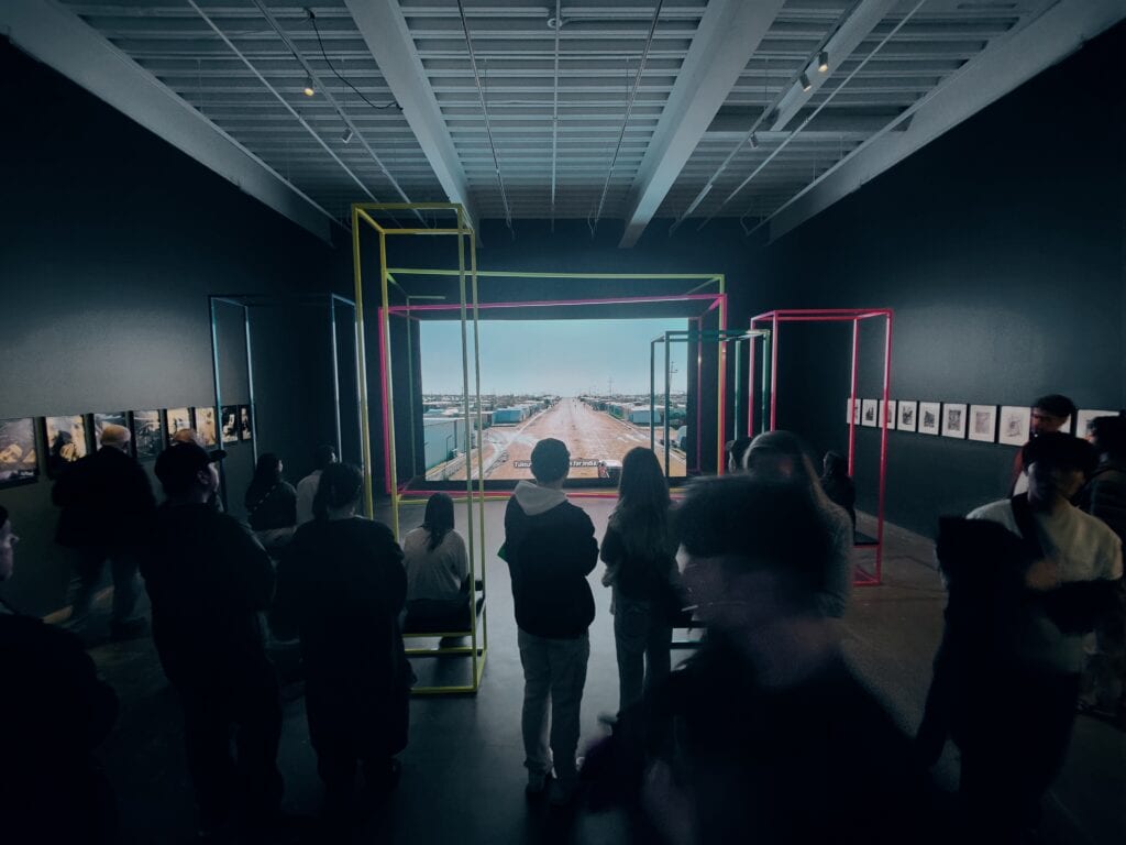 Photograph of dark gallery space in which figures stand in front of a video installation depicting a dirt road