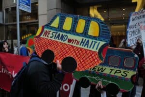 Protestors hold colorful signs shaped like buses. The words “We stand with Haiti” are written on one such sign.