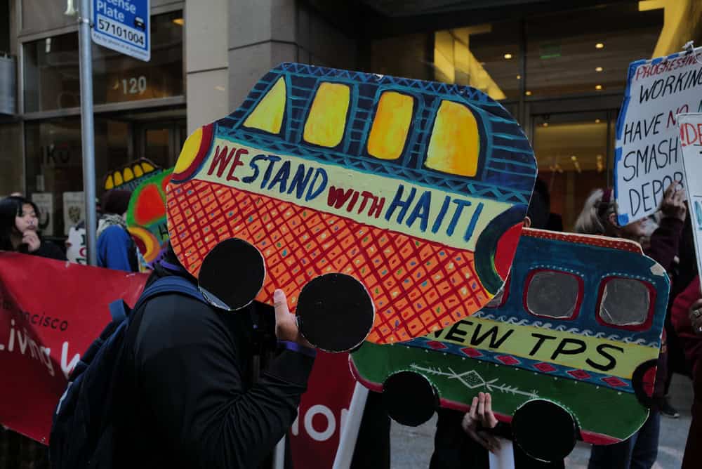 Protestors hold colorful signs shaped like buses. The words “We stand with Haiti” are written on one such sign.