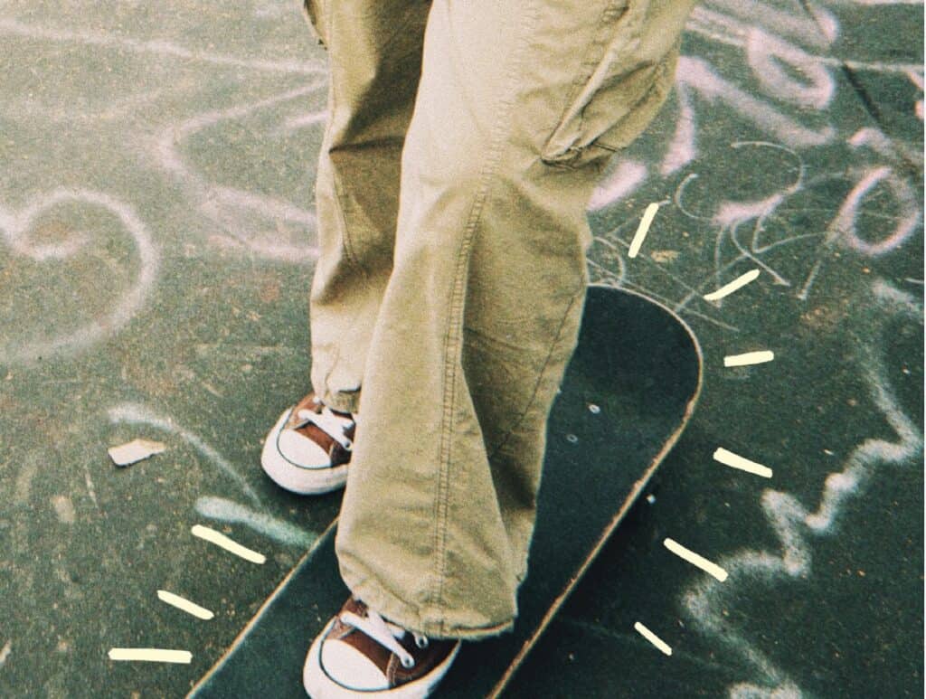 A film photo of a pair of legs in khaki pants, one foot on top of a skateboard. The ground beneath is covered in graffiti