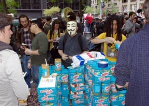 Photograph of an Occupy Wall Street protestor wearing a Guy Fawkes mask making a peanut butter sandwich from behind many stacked pallets of Skippy peanut butter