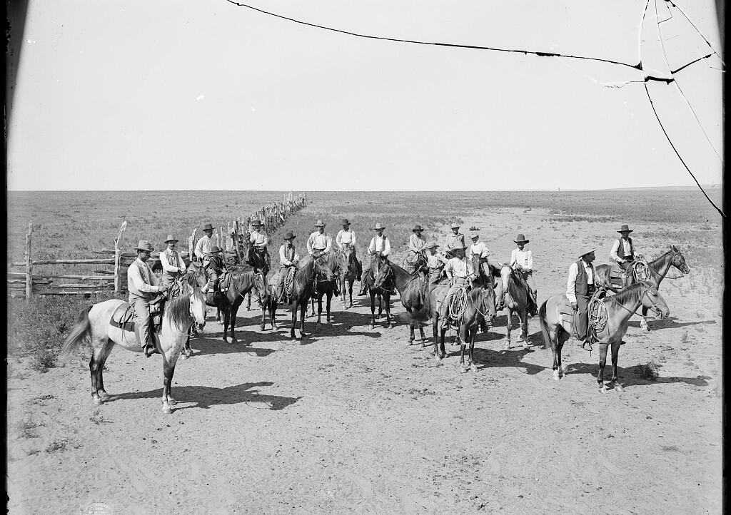 Black and white photograph of Texas cowboys lined up on horseback in an open field, a broken-down wooden fence to their right.