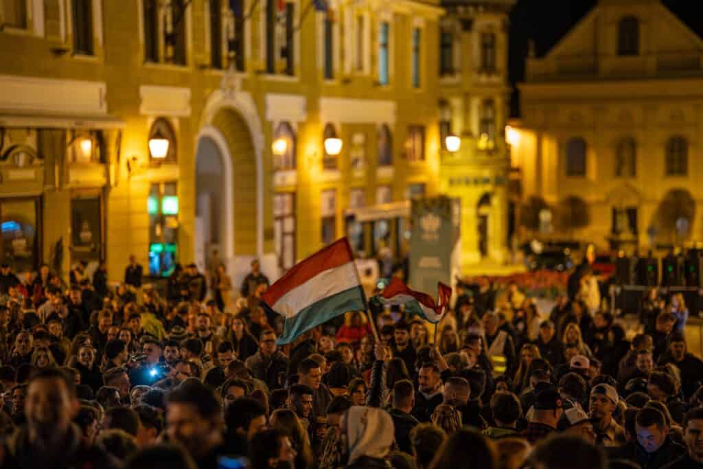 Photograph of a group of protestors gathered at night under city lights, waving Hungarian flags.